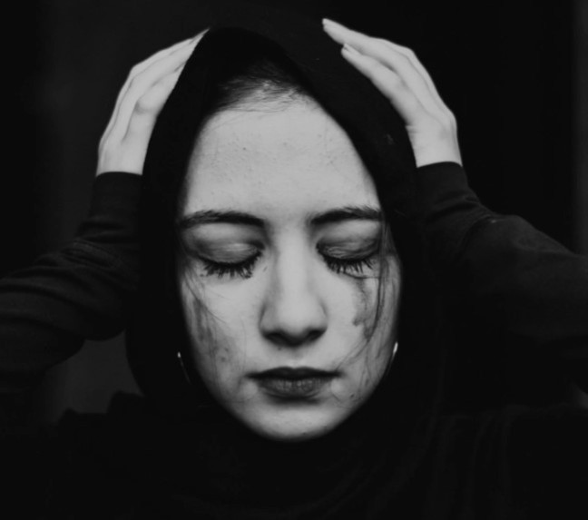 The image shows a woman whose face has black streaks of mascara on her cheeks. She wears a sad expression and has her hands placed on her head.